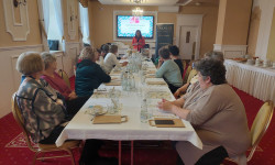 Sala konferencyjna. 20 kobiet siedzi przy długim stole. jedna osoba stoi i opowiada za nią wyświetla się prezentacja. Na stole  znajdują się kartki, małe i duże słoiki. 