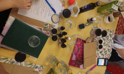 Stół nakryty żółtym obrusem a na nim fiolki, słoiczki, olejki. 