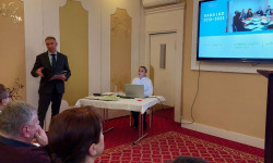 Konferencja LGD. Hrubieszów. W sali mężczyzna przemawia do zebranych osób. Za nim ekran z prezentacją. 