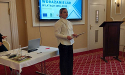 Konferencja LGD. Hrubieszów. W sali kobieta przemawia do zebranych osób. Za nią ekran z prezentacją. 
