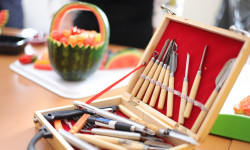 na stole arbuz wycięty w koszyk w którym są pokrojone owoce obok mała skrzynka z narzędziami służącymi do wycinania w owocach i warzywach  