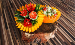 na kawałku płaskiego drewna dwie połówki dyni w jednej połowie w środku powycinane warzywa na kształt kwiatów w drugiej zielony sos 