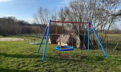 Na zdjęciu widoczne elementy placu zabaw, huśtawki, karuzela i boisko do siatkówki.