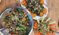 na drewnianym stole obok siebie  metalowa, okrągła miska z sałatką z marchewki i zielonych liści udekorowana kwiatami kończyny, owalny białawy talerz z pieczonymi plastrami batatów i smażonym szpinakiem udekorowany kwiatami stokrotki, szklana okrągła miska z ziołowym pesto udekorowana marchewką pociętoą w słupki i selerem 