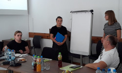 grupa ludzi przy stole i osoba prowadząca rozmawiają