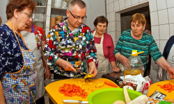w kuchni trzy kobiety i mężczyzna w fartuchach przy stole na którym pokrojone są warzywa z których mężczyzna przyrządza potrawę