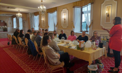 Sala konferencyjna. 20 kobiet siedzi przy długim stole. jedna osoba stoi i opowiada za nią wyświetla się prezentacja. Na stole  znajdują się kartki, małe i duże słoiki.