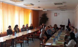 grupa ludzi przy stole i osoba prowadząca rozmawiają